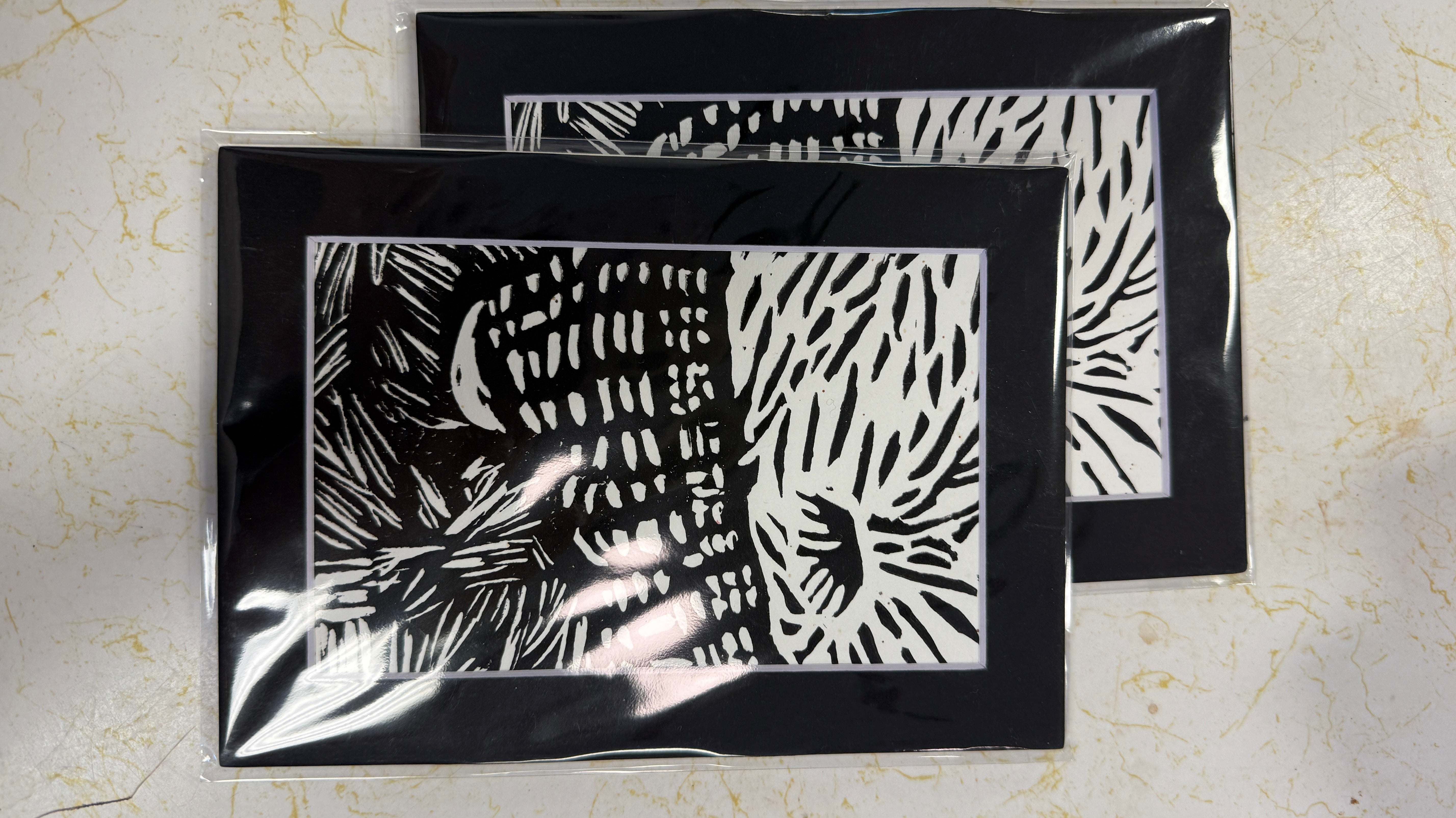 Two black-framed art prints with black-and-white linocut-style patterns, packaged in clear plastic sleeves and arranged on a