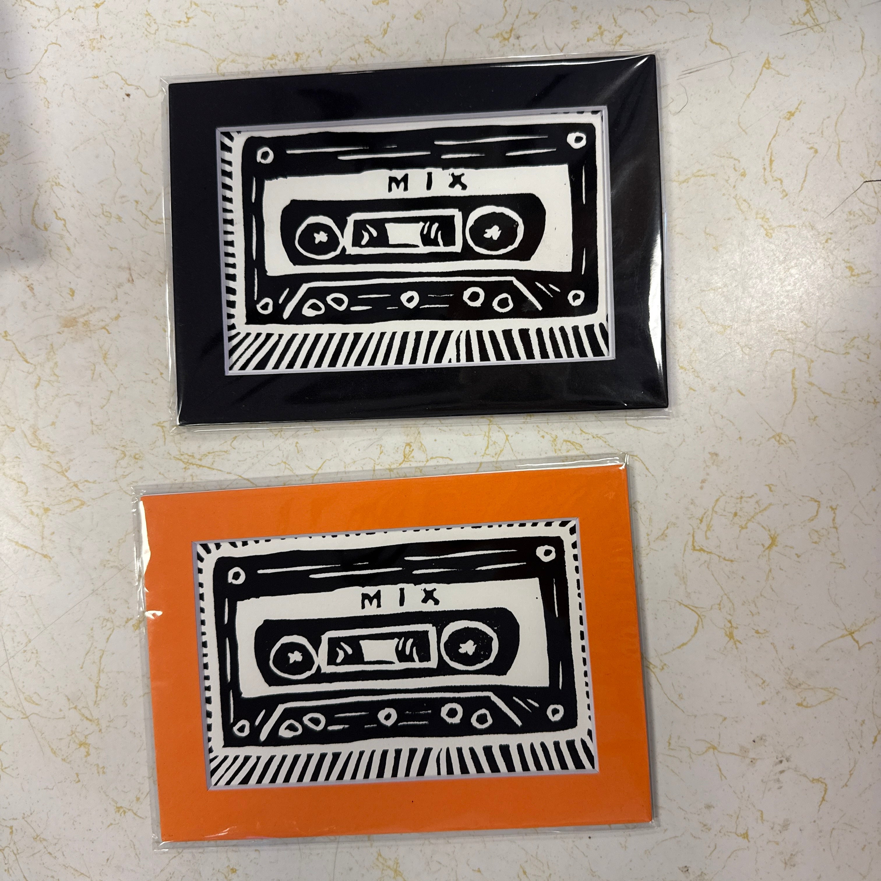 Two cassette tape art prints in clear plastic sleeves, one with an orange border and one with a black border, displayed on a