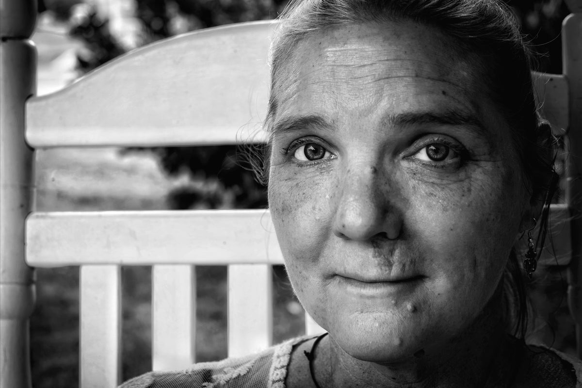 A black and white close-up portrait of a woman sitting in a wooden rocking chair, looking directly at the camera.