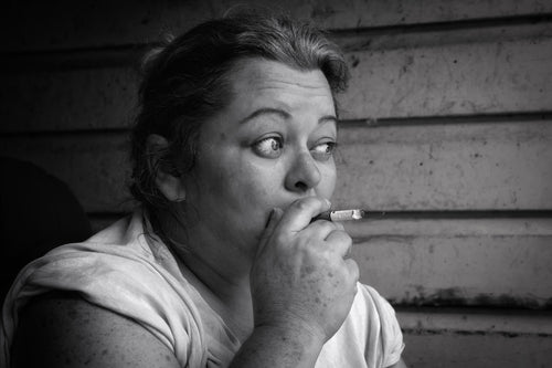 A black and white close-up portrait of a woman holding a cigarette near her mouth, wearing a light-colored top against a textured wooden wall.